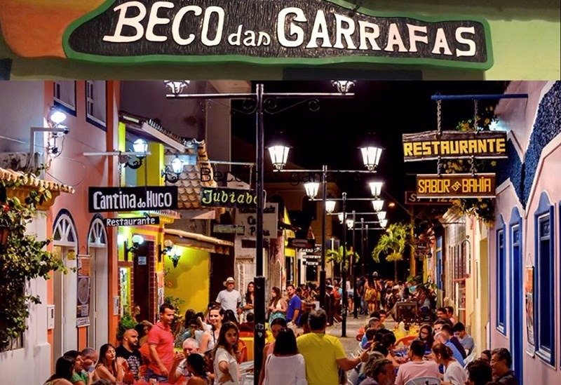 Vista noturna do charmoso Beco das Garrafas em Prado, Bahia, com mesas de restaurantes e iluminação aconchegante